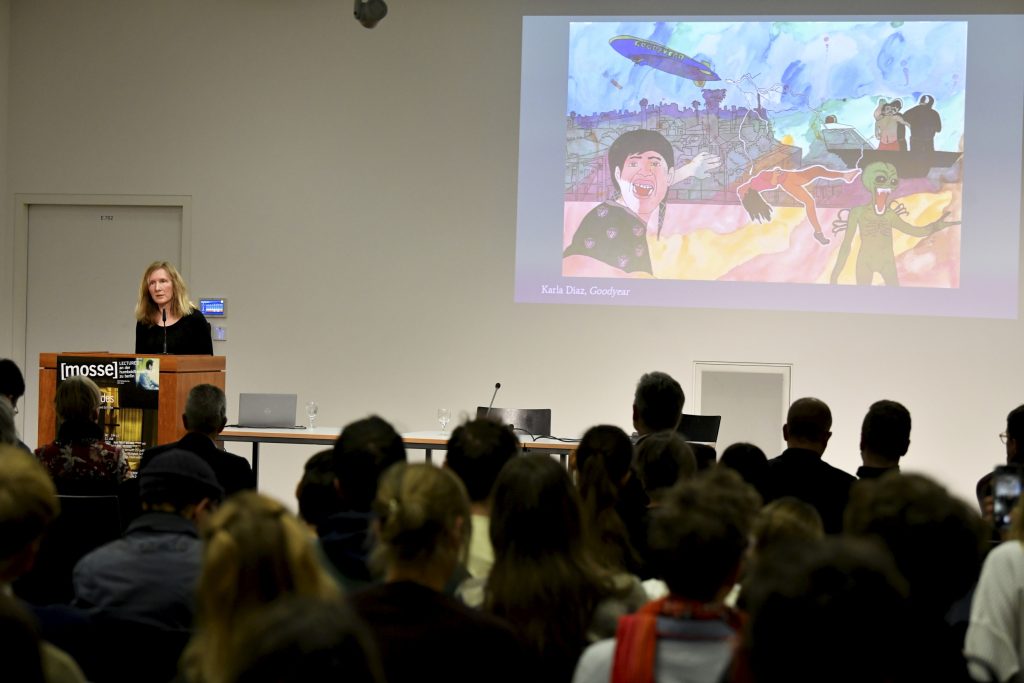 Samatha Harvey speaks at the podium, with an image projected behind her