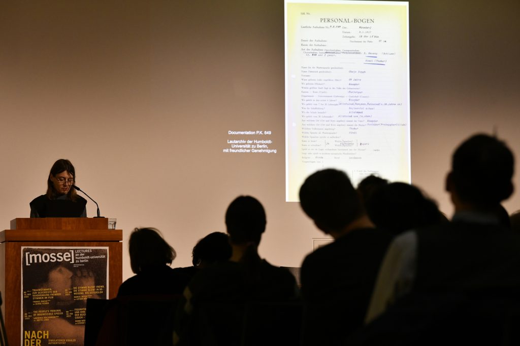 Britta Lange speaks at the podium, with a projected behind her