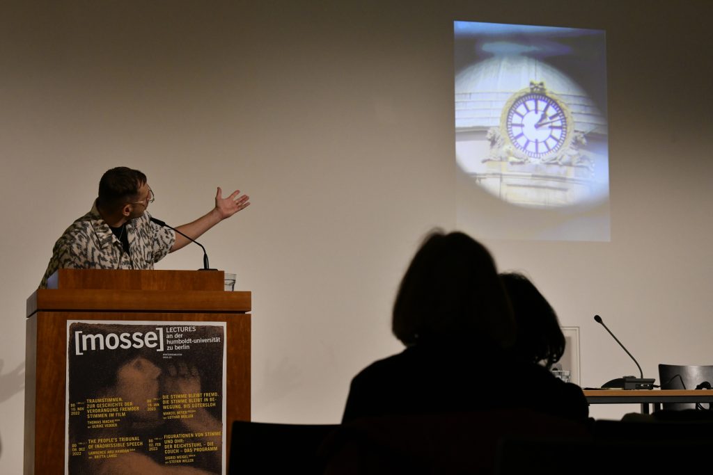 Hamdan delivering his lecture, with a slide in the background depicting a clock