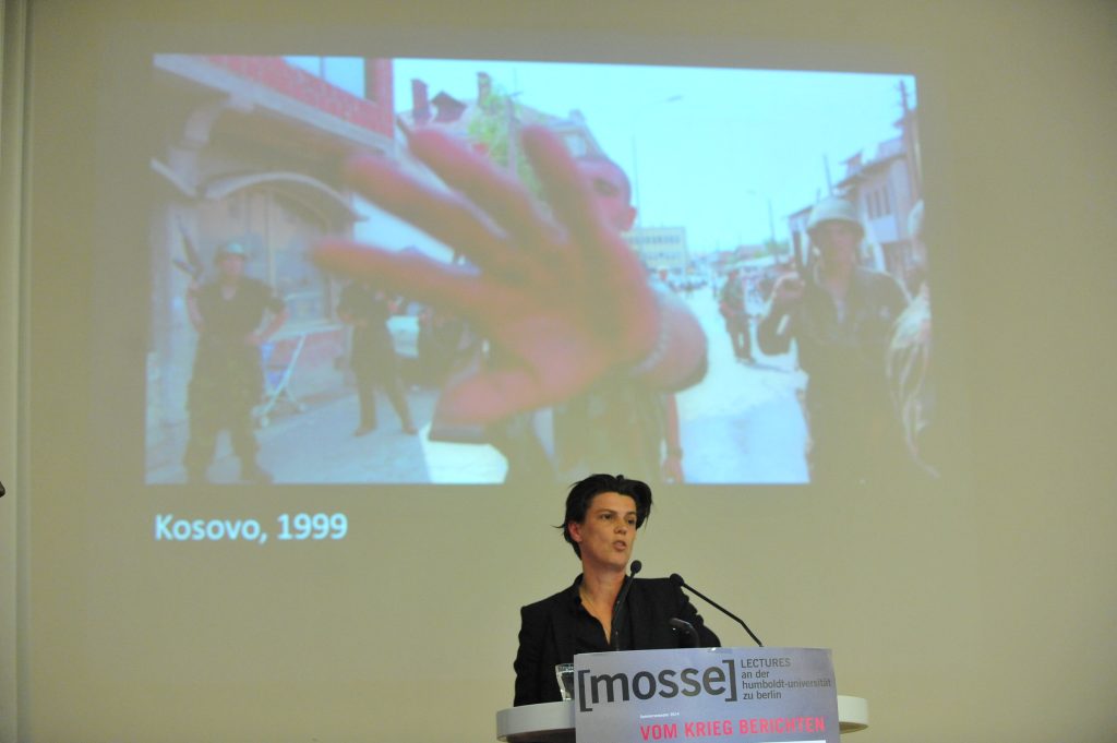 Emcke lectures from the podium, with photo in background