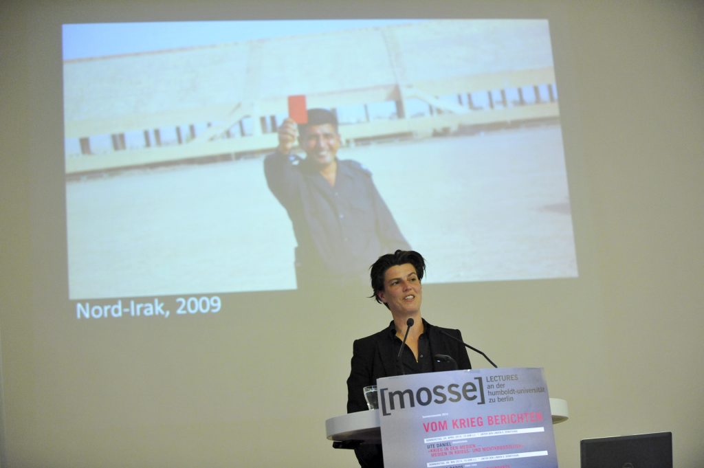 Emcke lectures from the podium, with photo in background